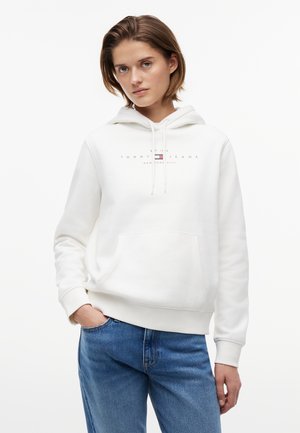 Młoda kobieta ubrana w biały kaptur Tommy Jeans z przednią kieszenią oraz niebieskie dżinsy, stojąca z jedną ręką w kieszeni.