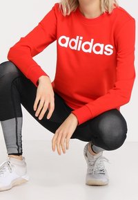 Felpa rossa con logo "adidas" bianco, abbinata a leggings neri testurizzati e scarpe da ginnastica grigio chiaro. Design semplice e vestibilità comoda.