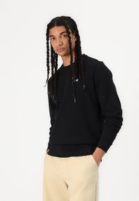 Ung person med lange fletninger har en sort sweatshirt og beige bukser på, stående med hænderne i lommerne foran en ensfarvet hvid baggrund.