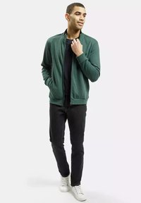 Zielona kurtka na zamek błyskawiczny noszona na czarnej koszulce, w połączeniu z ciemnymi dżinsami i białymi trampkami. Kurtka ma wysoki kołnierz i gładką fakturę.