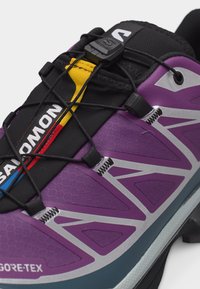 Artimas vaizdas į violetinį Salomon bėgimo batą su juoda greito raištelių sistema ir geltonais, raudonais bei mėlynais liežuvėlio akcentais.