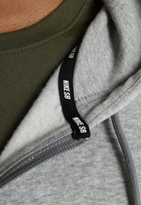 Felpa con zip grigia con una morbida fodera interna, caratterizzata da un'etichetta nera Nike SB sulla cerniera. Camicia verde oliva visibile sotto.