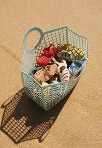 Šviesiai žydros spalvos pintinė su rankena, pripildyta įvairių spalvingų plastikinių šlepečių raudonos, rožinės, žalios, geltonos ir mėlynos spalvos, esančių ant smėlio paviršiaus.