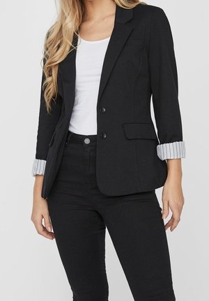 Femme portant un blazer noir avec des poignets rayés retroussés sur un haut blanc et un pantalon noir, montrée des épaules aux hanches.