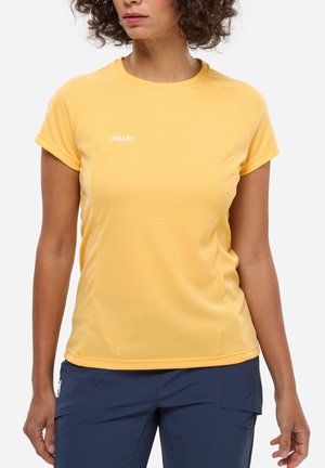 Femme portant un T-shirt jaune texturé à manches courtes et un pantalon bleu foncé, debout devant un fond blanc.