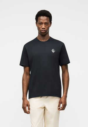 Jeune homme debout, face à l'avant, portant un t-shirt noir à manches courtes avec un petit logo sur la poitrine gauche et un pantalon beige.