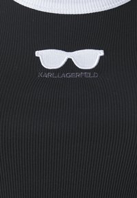 Svart ribbad tyg med en vit rund halsringning. Har en vit broderad solglasögonslogotyp och texten "KARL LAGERFELD" nedanför.