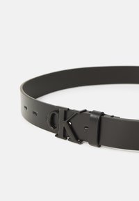 Svart läderbälte med slät yta, med en rektangulär spänne som har ett präglat "CK"-logotyp och flera justeringshål.