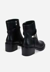 Botas de cuero negro hasta el tobillo con un tacón grueso, cremallera lateral y una correa metálica decorativa alrededor del tobillo. Textura suave y diseño elegante.