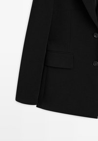 Blazer nero su misura con una superficie liscia, spalle strutturate, revers a punta, chiusura con un bottone singolo e tasca laterale.