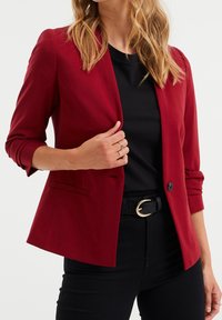 Blazer rojo con un corte entallado, cierre de un solo botón y mangas remangadas. Lleva puesto sobre una blusa negra, combinado con pantalones negros y un cinturón.