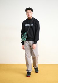 Sudadera negra con logo blanco, pantalones gris claro, bolso cruzado verde y zapatos negros con acentos rojos. De pie sobre un suelo amarillo.