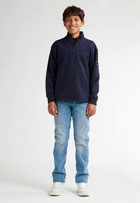 Hanorac albastru închis cu fermoar, mâneci lungi și guler tip stativ, asortat cu blugi deschis la culoare și adidași albi. Textură netedă.