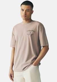 Lyserød bomulds-t-shirt med korte ærmer og rund hals, med et trykt logo der siger "Yellowstone Dutton Ranch" i mørk skrift.