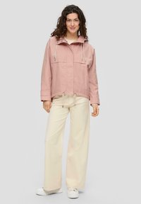 Lichtroze, gekruiste jas met elastische trekkoorden, twee grote voorkruimelpockets, gecombineerd met crèmekleurige wijde broek en witte platform schoenen.
