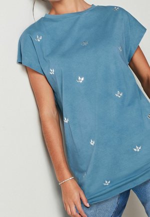 Personne portant une chemise bleue à manches courtes avec de petits motifs floraux blancs et un jean bleu clair, debout devant un fond clair uni.