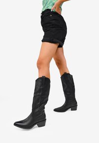 Stivali da cowboy in pelle nera con punta appuntita e tacco a blocco. Presentano cuciture decorative e linguette per facilitare l'indossare. Indossati con pantaloni corti neri arrotolati.