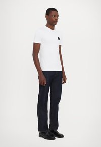 Hvid t-shirt med rund hals, der har et sort cirkulært logo på brystet, parret med mørkeblå lige ben jeans og sorte sko.