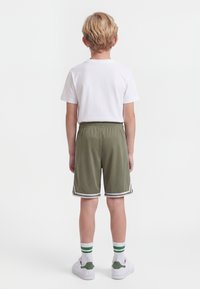 Niño con cabello rubio de espaldas, vistiendo una camiseta blanca, pantalones cortos verde oliva, calcetas blancas con rayas verdes y zapatillas blancas.