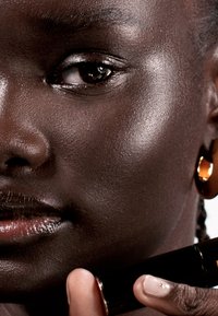 Primer plano de un rostro de piel oscura con piel suave, llevando un pendiente de aro dorado, sosteniendo un tubo de lápiz labial negro con uñas bien cuidadas.