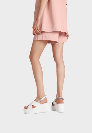 Gambe con pantaloncini rosa chiaro e camicia oversize, sandali bianchi con plateau con cinturini argento alla caviglia su sfondo grigio.