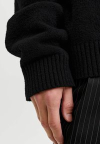 Mano descansando al lado del cuerpo, con un suéter negro texturizado con puño acanalado y pantalones negros con rayas finas.