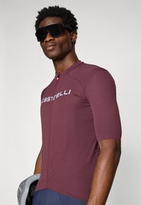 Maillot de cyclisme bordeaux à manches courtes avec une fermeture éclair devant, arborant un logo blanc sur la poitrine et une coupe aérodynamique. Porté avec des lunettes de soleil noires.