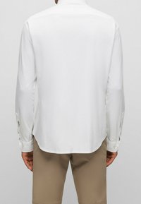 Camicia bianca a maniche lunghe con colletto, caratterizzata da un orlo curvo e polsini con bottoni, realizzata in tessuto morbido. Nessun motivo o dettaglio visibile.