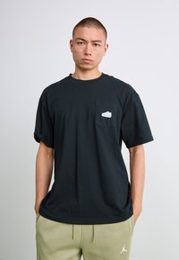 Mand med kort hår iført en ensfarvet sort T-shirt med et lille sneakersmærke på brystlommen og lyse olivengrønne sweatpants, der står op ad en hvid væg.