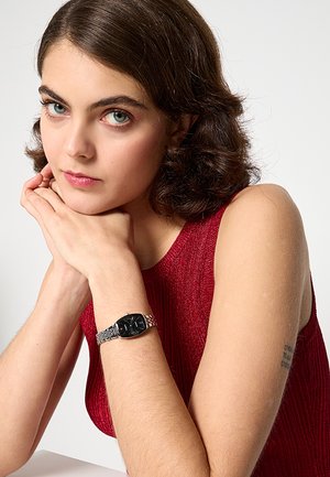 Mladá osoba s krátkymi hnedými vlasmi a modrými očami, oblečená v červenom tielku bez rukávov, opierajúca si bradu o ruky a ukazujúca strieborné hodinky na zápästí.