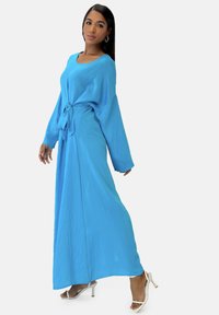Lyseblå maksimalangærmet kjole med bindebånd i taljen og flydende silhuet. Fremstillet af blødt stof med mat tekstur. Sko med høje hæle er synlige.