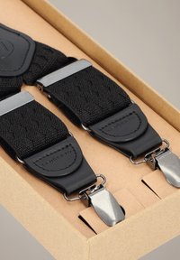 Czarne szelki z tkaniny o teksturowanym splocie, z elementami ze skóry i metalowymi klipsami, zaprezentowane w kartonowym pudełku.