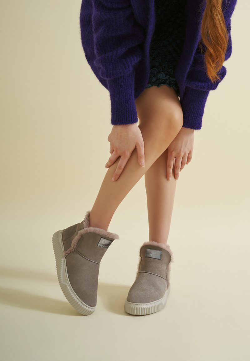 Stivali alla caviglia in suede grigio con foderatura pelosa rosa, suola in gomma testurizzata e un patch con logo argentato. Modella seduta, ginocchio piegato, che indossa un maglione viola.