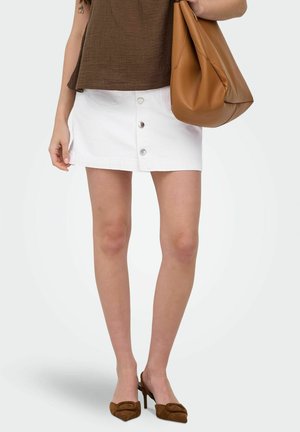 Žena v bielej gombíkovej mini sukni, hnedom tielku bez rukávov, hnedých lodičkách na vysokom opätku, nesúca veľkú hnedú koženú tašku na ramene.