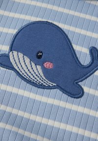 Sininen raidallinen kangas, jossa on brodeerattu sininen valaiden kuva valkoisella vatsalla, mustilla silmillä ja vaaleanpunaisella poskipunalla poskessa.