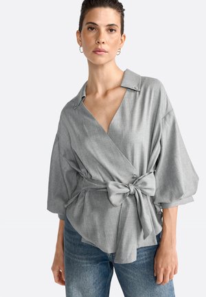 Gray wrap blouse with a notched collar and short, wide sleeves. Includes a tied waist and subtle texture. Worn with light blue jeans.