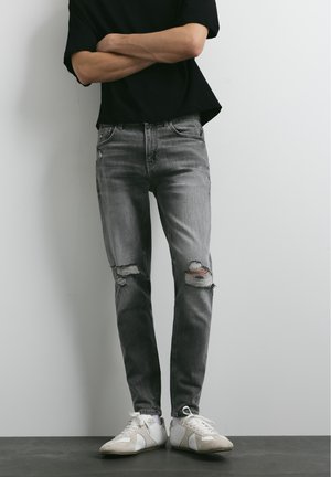 Personne portant un jean gris déchiré, des baskets blanches et un t-shirt noir à manches courtes, bras croisés, debout contre un mur uni.