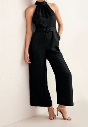 Frau trägt einen ärmellosen schwarzen Halfter-Jumpsuit mit tailliertem Gürtel und weitem Bein, kombiniert mit beigen High-Heel-Sandalen.