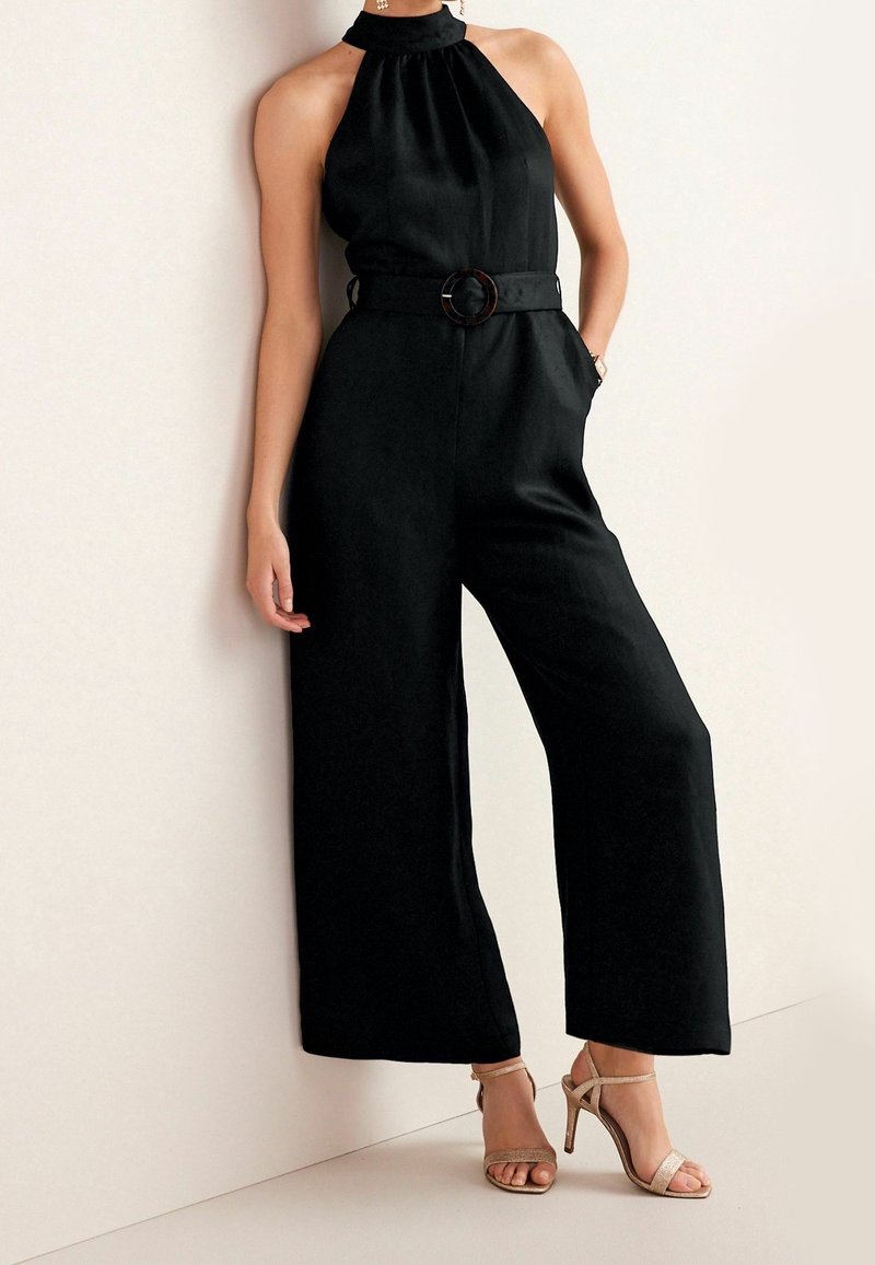 Femme portant une combinaison noire sans manches à col halter avec une taille ceinturée et un pantalon à jambes larges, associée à des sandales à talons hauts beiges.