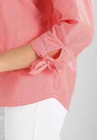 Blouse rayée rouge et blanche avec des manches retroussables à nouer. Le tissu est léger avec une texture lisse. L'ourlet inférieur est légèrement incurvé.