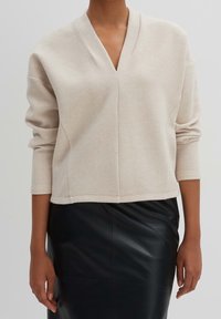 Maglione lavorato a maglia beige con scollatura a V, maniche lunghe e silhouette corta, abbinato a una gonna a matita in pelle nera. Tessuto morbido e testurizzato.