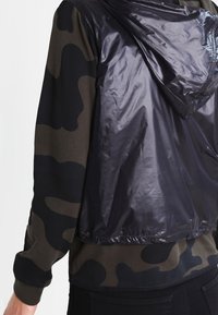 Persona con un chaleco negro brillante con capucha sobre una camiseta de manga larga con patrón de camuflaje, con pantalones negros visibles abajo.