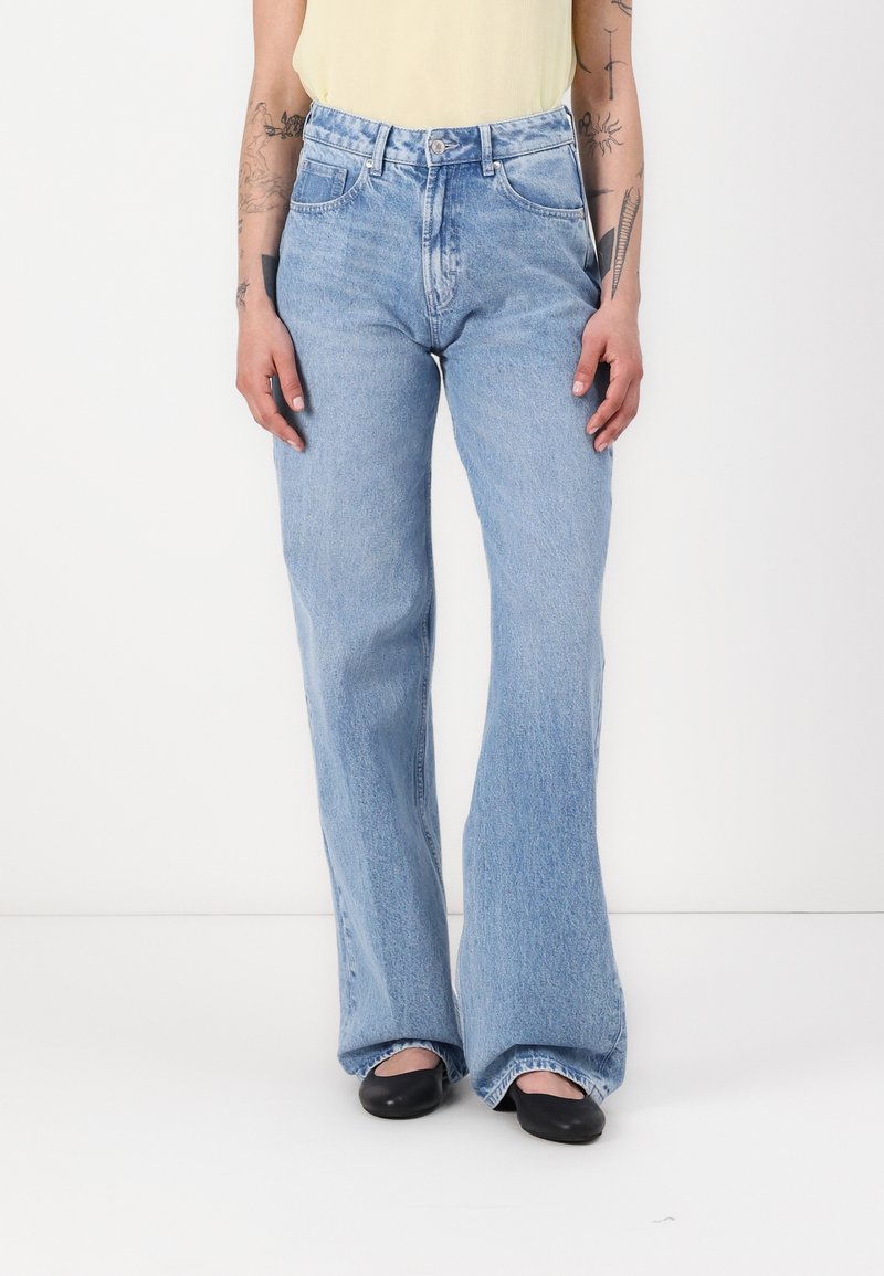 Un jean flare bleu clair présente une taille haute, un design à cinq poches et une texture en denim douce. Porté avec un haut jaune et des ballerines noires.