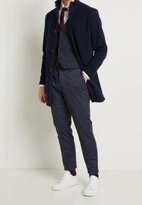 Hombre vestido con un abrigo azul marino, pantalones a cuadros azul oscuro, zapatillas blancas, calcetines burdeos, camisa blanca, chaleco azul marino y corbata burdeos, de pie contra una pared blanca.
