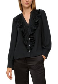 Frau in einer schwarzen, langärmeligen Bluse mit großen, gefältelten Rüschen an der Vorderseite und schwarzen Satin-Hosen, die eine Hand in der Tasche hat.