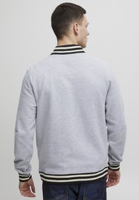 Grå hettegenser med en svart-hvitt stripet krage og mansjetter, med lange ermer og ribbestrikket kant. Enkel design, ingen synlige mønstre.