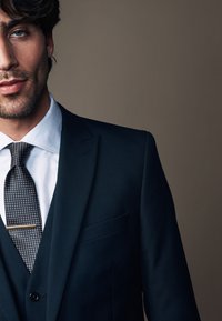 Dunkelblaues Anzugjackett mit Revers, kombiniert mit einem hellblauen Hemd und einer gemusterten Krawatte, die mit einer goldenen Krawattennadel fixiert ist. Strukturierte Stoffdetails sind sichtbar.