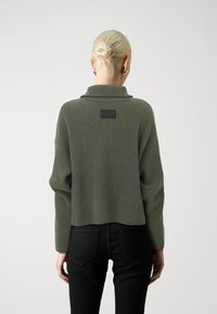 Olivgrüner, gerippter Pullover mit hohem Kragen, hinten mit einem schwarzen Etikett. Kombiniert mit schwarzen, eng anliegenden Jeans.