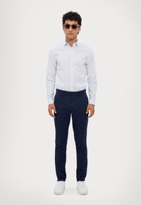 Lyseblå skjorte med knapper, marineblå slim-fit bukser, hvite sneakers og mørke solbriller; glatt stoff og skreddersydd design.