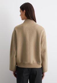 Beige, struktureret, oversized sweater med ribbet krave og kant. Har sænkede skuldersømme og en glat overflade, stylet med mørke bukser.